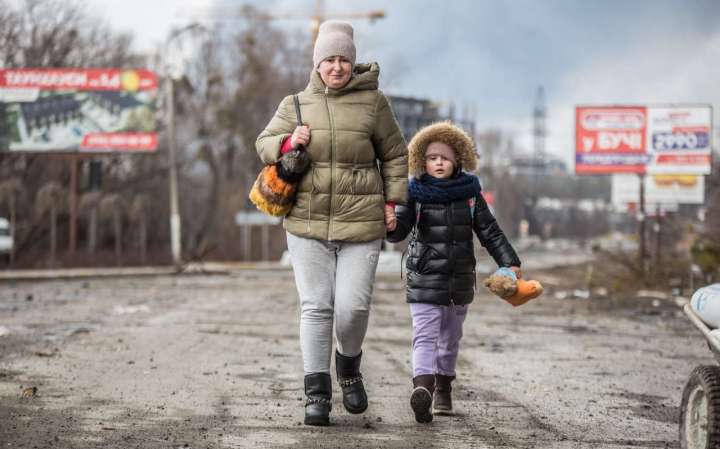 Ukrainalaiset äiti ja lapsi kävelevät käsikädessä sodan runtelemassa kaupungissa. Äidin kasvoilla on huolestunut ilme.