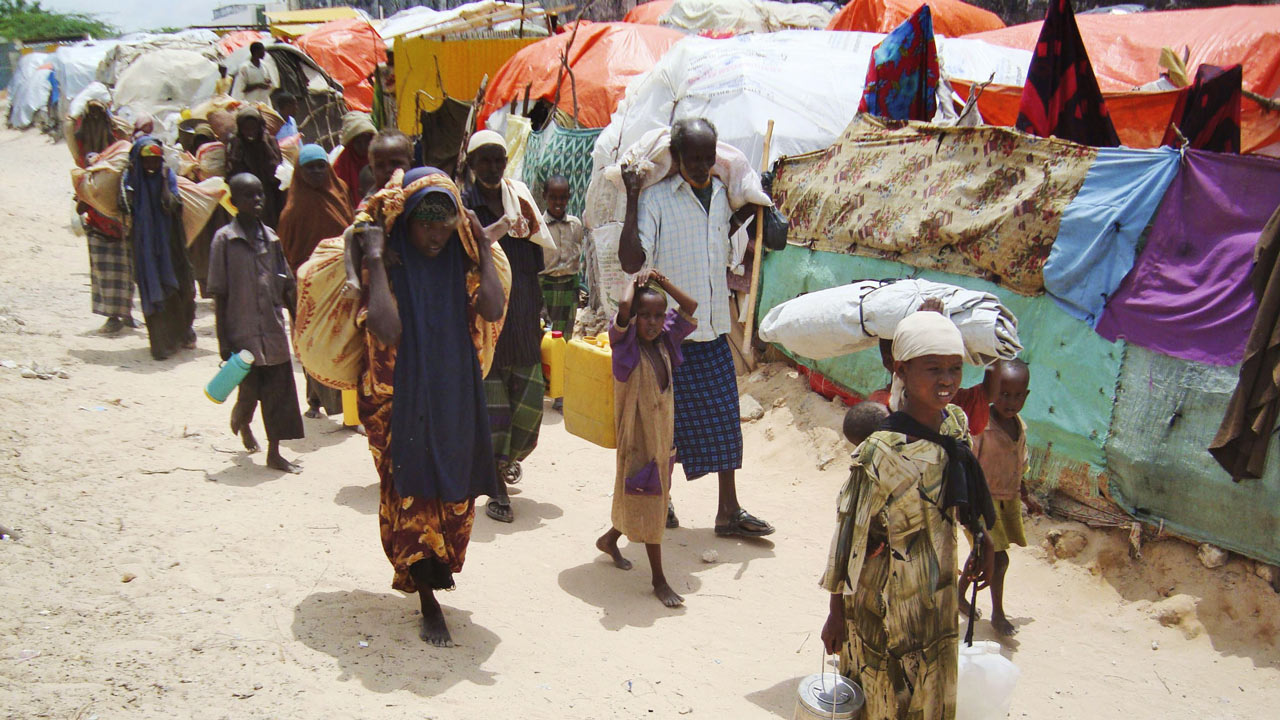 Perhekunta eri ikäisiä ihmisiä kuvattuna kävelemässä nyytteineen pakolaisleirin edustalla