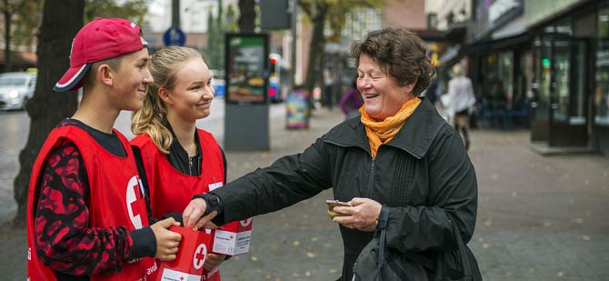 Leende flicka och pojke samlar pengar för Röda Korset på gatan, och en leende äldre kvinna tar del i insamlingen